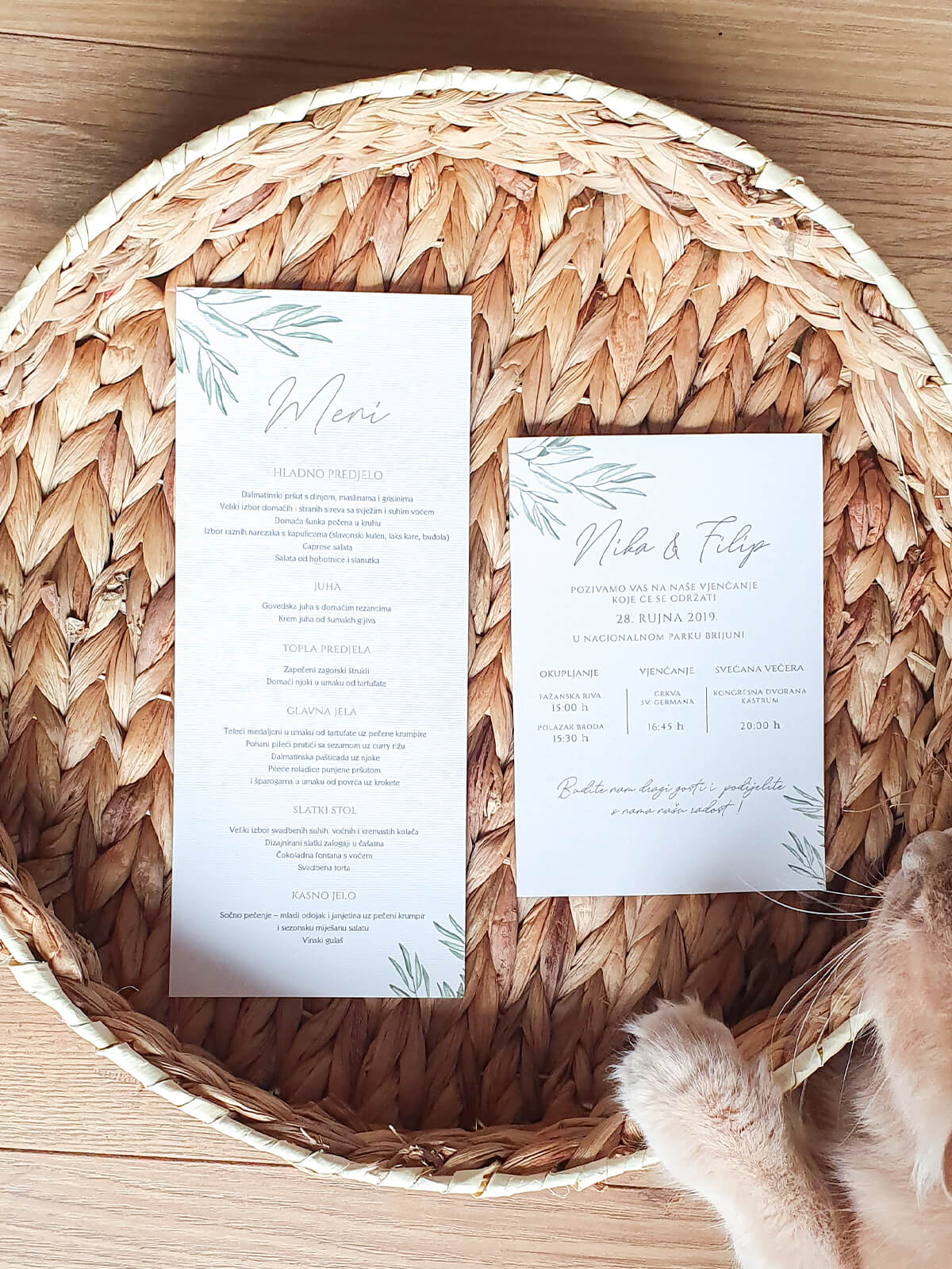 Meni i pozivnica za vjenčanje s dizajnom eukaliptusa postavljeni na pleteni tanjur s vidljivim mačjim šapama koji ih pridržavaju