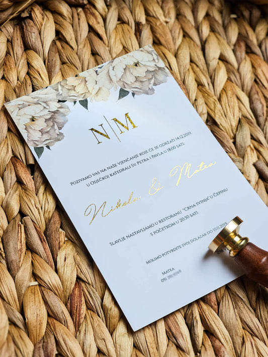 Pozivnica za vjenčanje sa zlatnim inicijalima i cvjetnim detaljima na gornjem dijelu, prezentirana na pletenoj podlozi